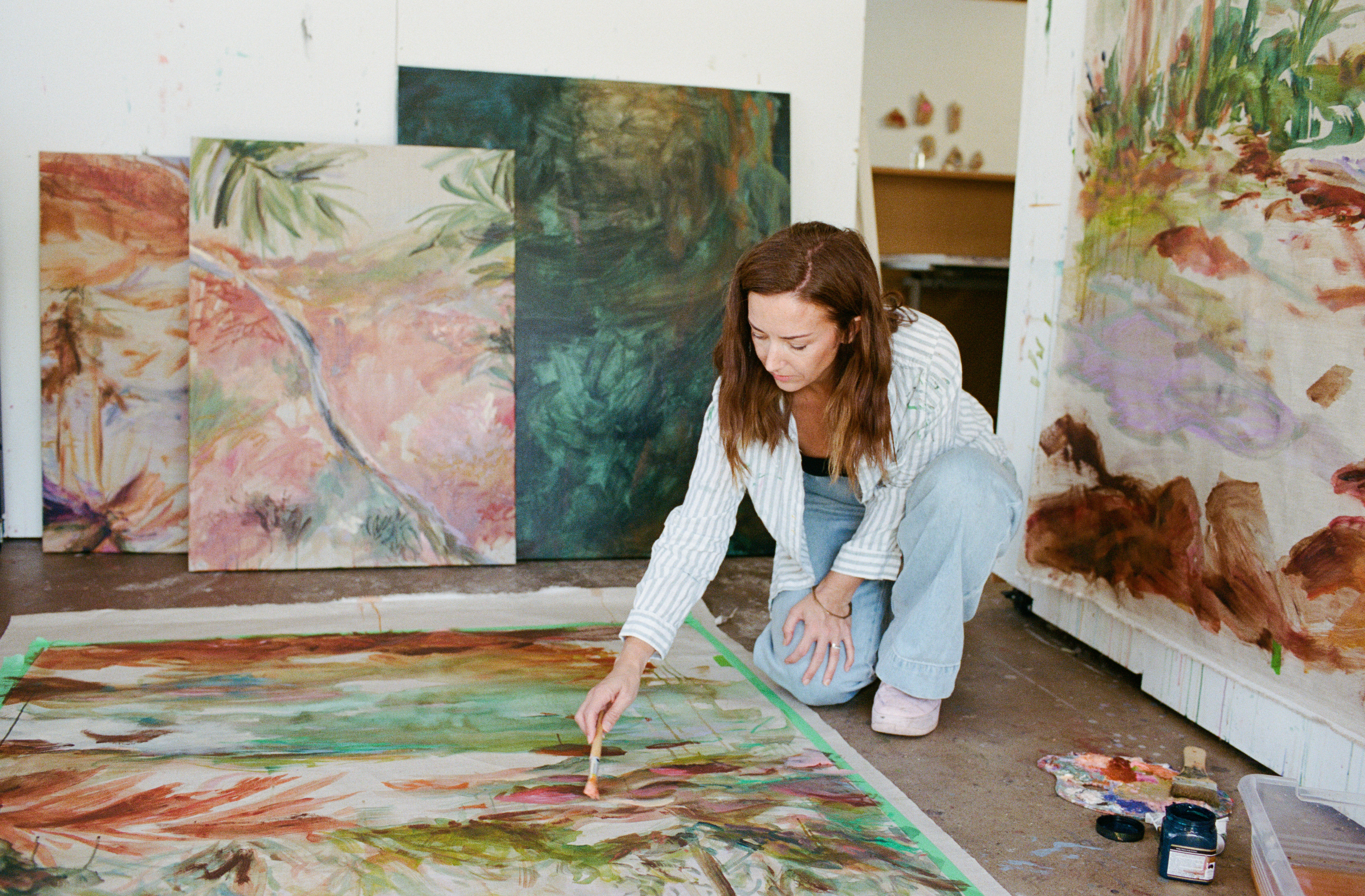 An artist crouches in an art studio, painting on a large canvas laid out on the floor. Several large colorful paintings are leaning against the walls, and paint supplies sit on the floor nearby.